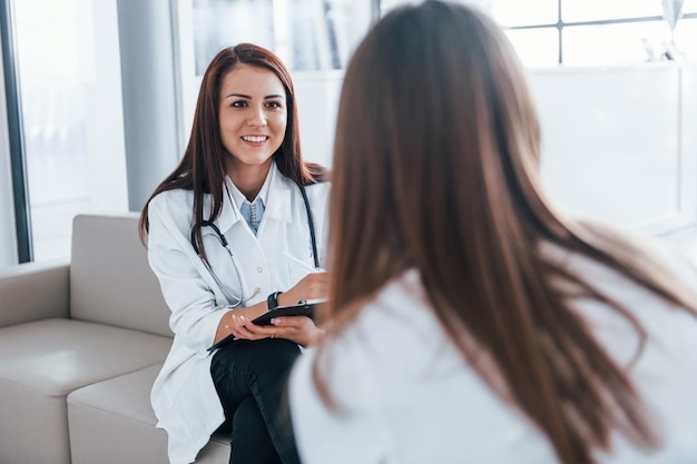 Zitten en praten Jonge vrouw heeft een bezoek aan een vrouwelijke arts in een moderne kliniek