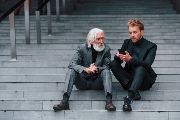 Zittend met telefoon Jonge man met senior man in elegante kleding is buiten samen Conceptie van zaken