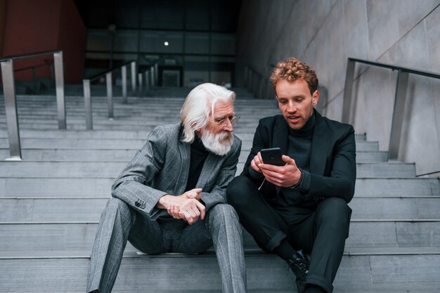 Zittend met telefoon Jonge man met senior man in elegante kleding is buiten samen Conceptie van zaken