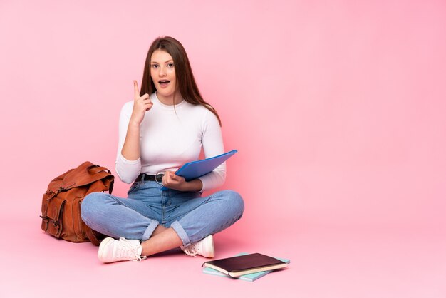 Zitting van het de studentenmeisje van de tiener de kaukasische die op de vloer op roze muur wordt geïsoleerd denkend een idee dat de vinger benadrukt