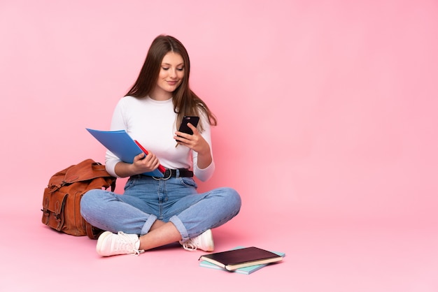 Zitting van het de studentenmeisje van de tiener de Kaukasische die op de vloer op roze wordt geïsoleerd die een bericht met mobiel verzenden