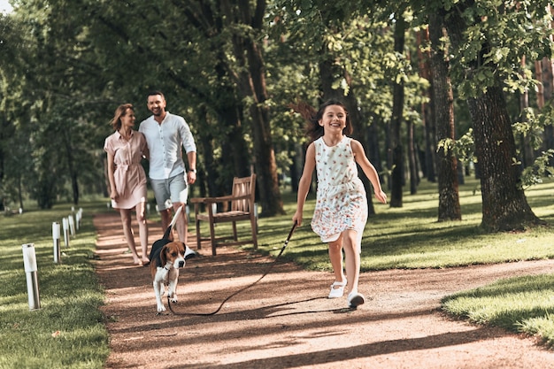 Zo fijn om samen te zijn! Volledige lengte van schattig klein meisje met hond die rent en glimlacht tijdens het wandelen met haar ouders in het park