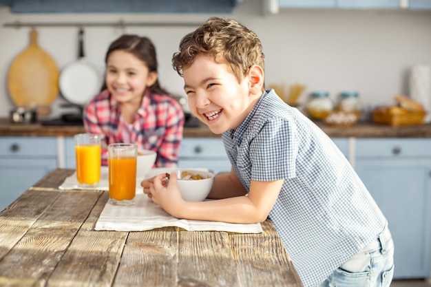 Zo vrolijk. Knap opgetogen kleine donkerharige jongen lachen en gezond ontbijten met zijn zus en het meisje lachend in de