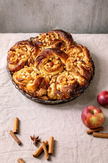 Zoete appel plakkerige kaneelbroodjes in ronde bakplaat met tuinappels en kaneelstokjes op tafel met grijs linnen tafelkleed. Traditionele thuisbakkerij