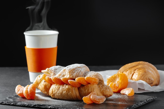 Zoete croissantsandwich met mandarijnen kopje koffie met poedersuiker op een donkere baki als achtergrond