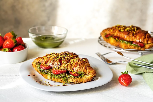 Zoete croissantsandwiches met pistachecrème, aardbeien en karameldip op witte houten tafel en grijze achtergrond