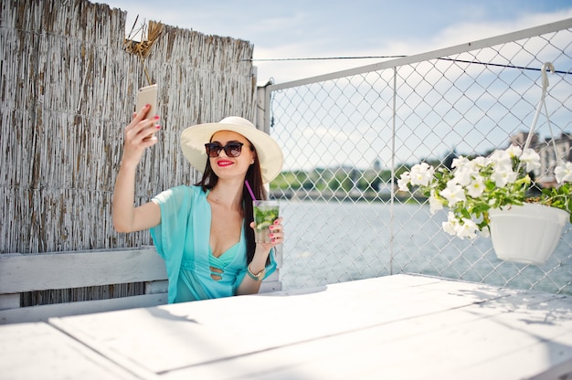 Zoete jonge dame die hoed en zonnebril met een cocktail draagt die haar smartphone gebruikt terwijl het zitten door het meer.