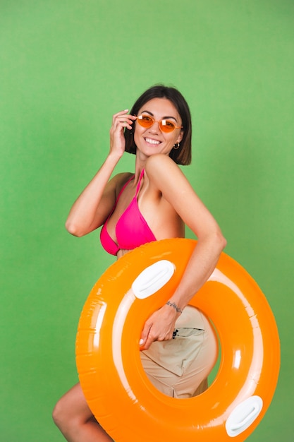 Zomer fit sportieve vrouw in roze bikini en fel oranje opblaasbare ring rond en zonnebril op groen, blij vrolijk opgewonden blij positief