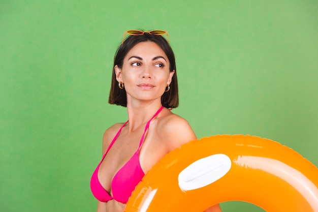 Zomer fit sportieve vrouw in roze bikini en fel oranje opblaasbare ring rond en zonnebril op groen, blij vrolijk opgewonden blij positief