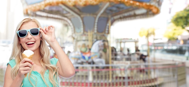 Foto zomer, junkfood en mensenconcept - jonge vrouw of tienermeisje in zonnebril die ijs eet over carrousel bij pretparkachtergrond