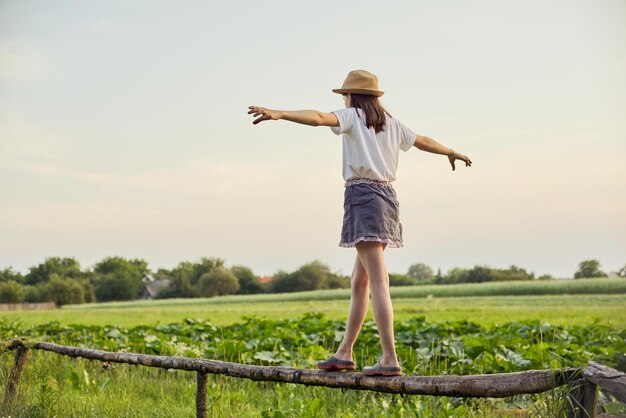 Zomer, natuur, geluk, jeugdconcept. Meisje in een hoed lopen op een landelijke houten hek, prachtige zonsondergang landschap-achtergrond.