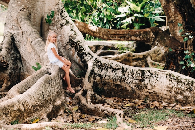 Zomer portret van een gelukkig klein meisje op het eiland mauritius zittend op een enorme boom. een meisje zit op een grote boom in de botanische tuin van het eiland mauritius.