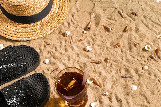 Zomer strandkleding, slippers, hoed, koud drankje in een glas en schelpen op zandstrand.