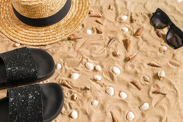 Zomer strandkleding, slippers, hoed, zonnebril en schelpen op zandstrand.