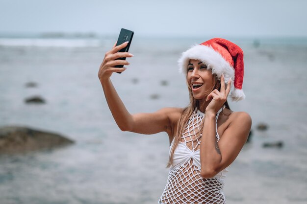 Zomer strandvakantie meisje in kerstmuts die leuke mobiele selfie foto maakt met smartphone. Meisje dragen witte zwembroek poseren voor selfie. Trips naar warme bestemmingen. Phuket. Thailand.