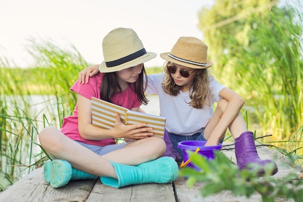 Zomer, vakantie, natuur, levensstijl, vrije tijd voor kinderen. Kinderen, twee meisjes zittend op een houten pier in het rietmeer, pratend en lezend notitieboekje met interesse