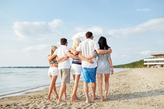 zomer, vakantie, zee, toerisme en mensen concept - groep lachende vrienden knuffelen en wandelen op het strand vanaf de achterkant