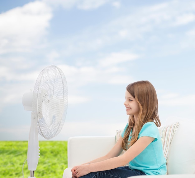 Zomer, weer en uitrusting concept - glimlachend klein meisje zittend op de bank met grote ventilator thuis