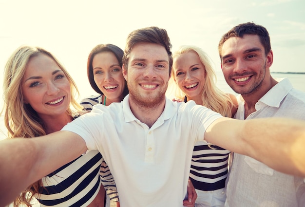 zomer, zee, toerisme, technologie en mensen concept - groep lachende vrienden met camera op strand fotograferen en selfie nemen