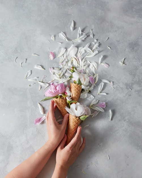 Zomerbloemen - verse zachte roze en witte pioenroos in een wafelkegels met vrouwelijke handen, bloemblaadjes op een grijze marmeren tafel. Plaats voor tekst, bovenaanzicht.