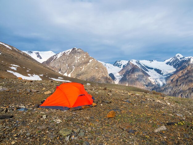 Zomerkamperen in de bergen Helder alpenlandschap met levendige oranje tent op zeer grote hoogte met uitzicht op hoge berg en grote gletsjer in donkere wolken Geweldig berglandschap met tent