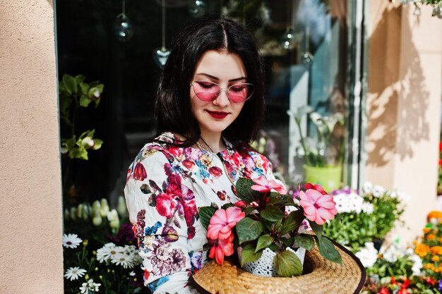 Zomerportret van brunette meisje in roze bril en hoed tegen bloemenwinkel