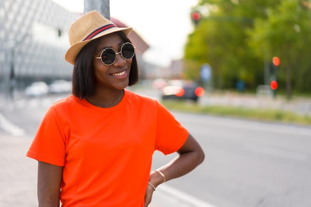 Zomerpret Jonge zwarte vrouw met zonnebril en hoed met oranje t-shirt die glimlacht in de levensstijlfoto's van de stad