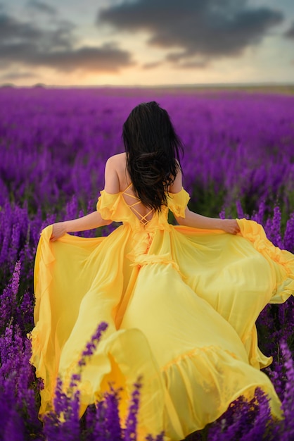 Zomerstemming. Een vrouw in een luxe gele jurk loopt langs een paars bloeiend veld met haar rug naar de camera.