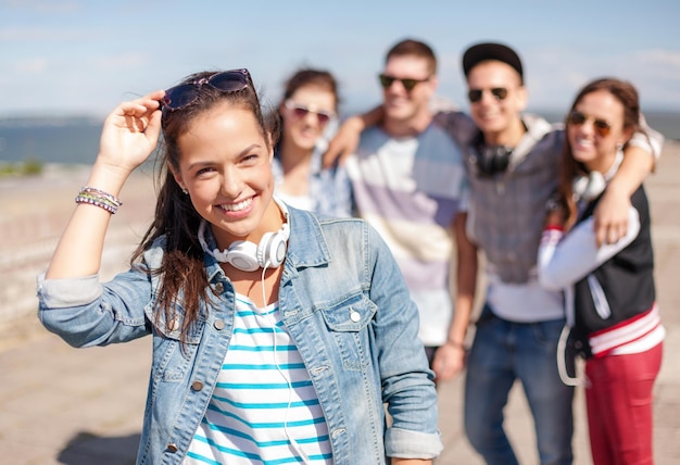 zomervakantie en tienerconcept - tienermeisje met zonnebril en koptelefoon die buiten met vrienden rondhangt