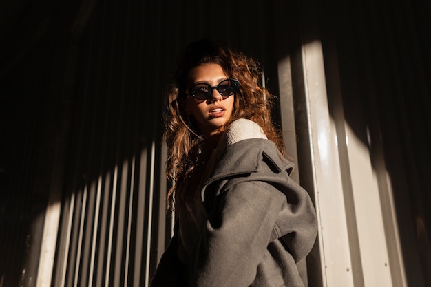 Zonlichtportret van een mooi modieus meisje met krullend haar in modekleding met een stijlvolle zonnebril en een vintage jas op straat in de buurt van een metalen muur in de schaduw