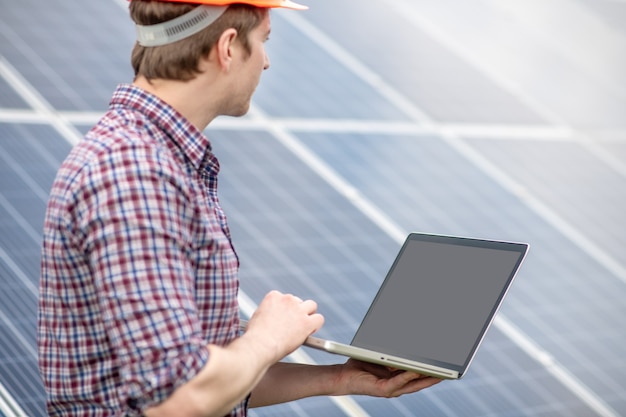 Zonne energie. Man in geruit hemd met laptop in zijn handen kijkend naar het oppervlak van de zonnebatterij die ernaast staat