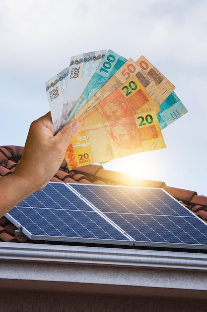 Zonne-energieeconomie, Braziliaans geld voor het fotovoltaïsche paneel op een dak, zonsondergangachtergrond. Ruimte voor tekst.