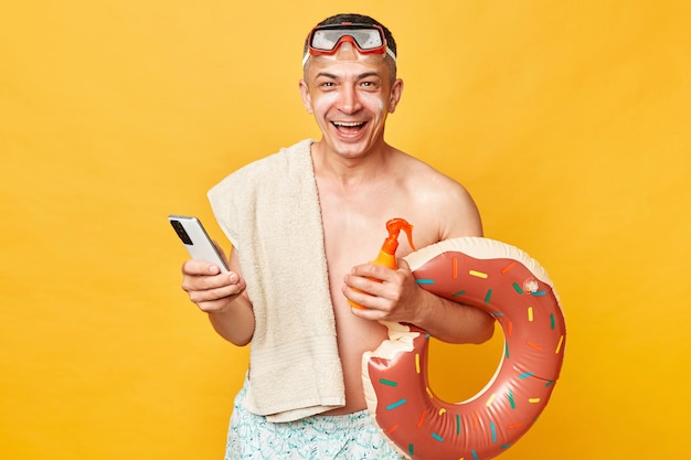 Zonnebaden bij het zwembad van het resort Gelukkig opgewonden volwassen man met korte zwembroek zwembroek snorkelbril met donut rubberen ring en tas geïsoleerd op gele achtergrond met behulp van telefoon browsen op internet