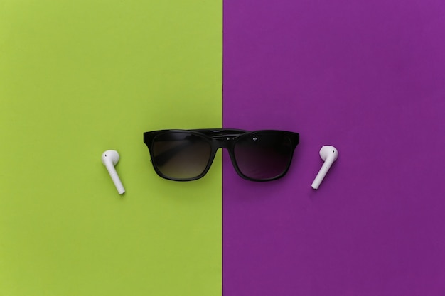 Zonnebrillen en draadloze koptelefoons op een paarsgroene achtergrond.