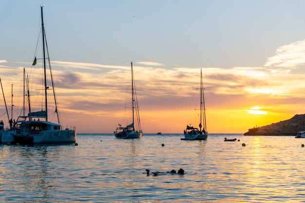 Zonsondergang op Ibiza. Twee jonge vrouwen zwemmen en verschillende schepen voor anker met mensen kijken naar de spectaculaire zonsondergang boven de oceaan met zijn dramatische kleurveranderingen