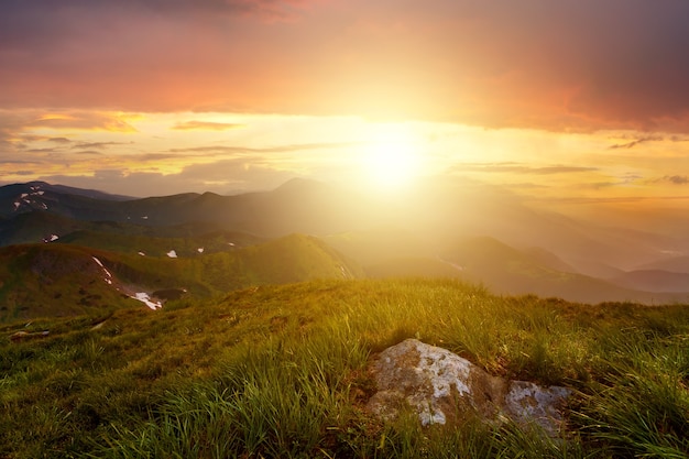 Zonsonderganglandschap met groene grasweide, hoge toppen en mistige vallei onder levendige kleurrijke avondlucht in rotsachtige bergen.