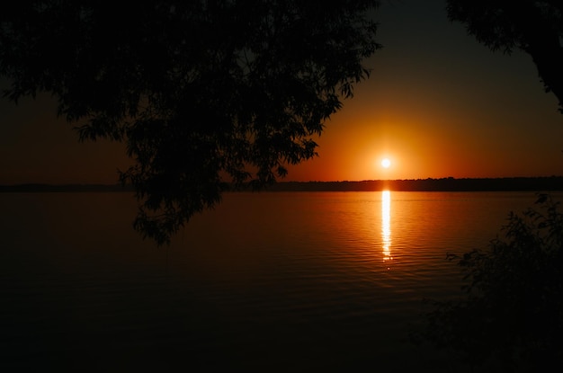 Zonsonderganglandschap op meer Ondergaande zon en reflectie op watersilhouetten van bomen