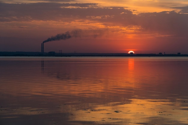 Zonsondergangmening van rokende industriële pijpen op een horizon over de rivier