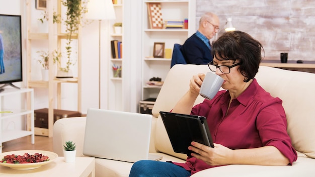 Zoom in op een foto van een oudere vrouw die een tablet gebruikt die op een bank zit. senior man leest een boek op de achtergrond.