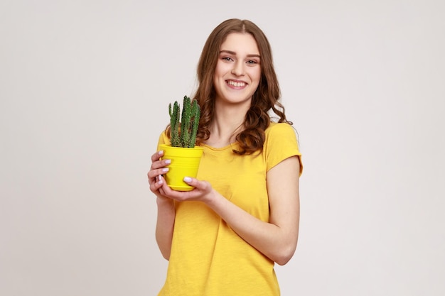 Zorg voor potplant portret van een jonge vrouw in gele T-shirt met bloempot met cactus kijkend naar camera met brede glimlach Indoor studio opname geïsoleerd op een grijze achtergrond