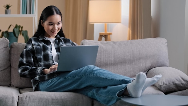 Zorgeloos Aziatisch meisje komt de woonkamer binnen, gaat op een comfortabele bank zitten en neemt een laptop met een glimlach horloges