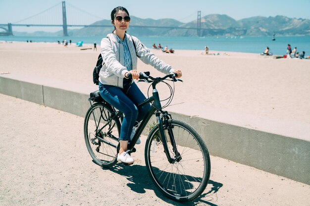 Zorgeloos aziatisch meisje met een zonnebril heeft zomervakantieplezier door met de fiets langs het strand in san francisco, californië, vs met zonneschijn te fietsen.