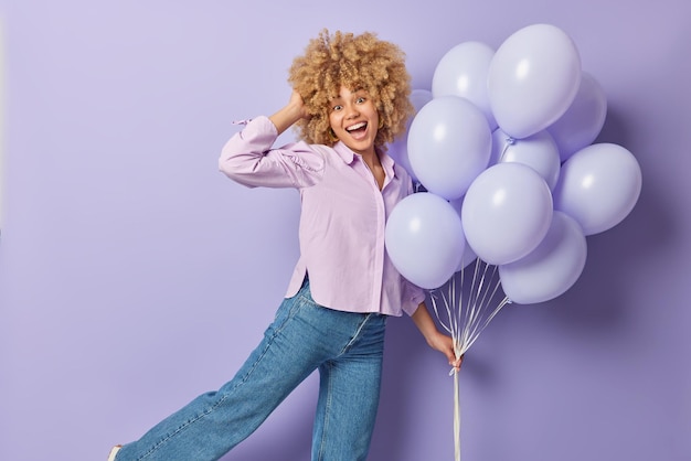 Zorgeloos blij krullend haired vrouw dansen met bos van opgeblazen ballonnen draagt shirt en spijkerbroek geniet van party time geïsoleerd over paarse achtergrond Mensen viering en speciale gelegenheid concept