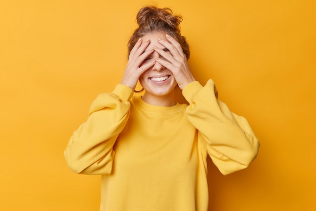 Zorgeloos blije vrouw bedekt ogen met handen wacht op verrassing stads vrolijk heeft haar in knot gekamd draagt casual trui geïsoleerd op gele achtergrond speelt verstoppertje anticipeert op iets goeds