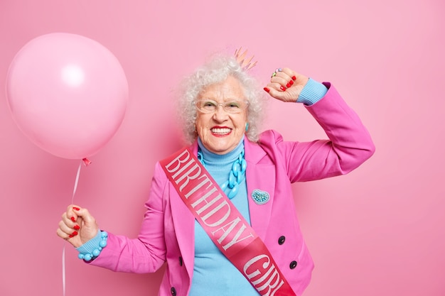 Foto zorgeloos grijsharige gerimpelde vrouw danst zorgeloos glimlacht positief gekleed in feestelijke kleding draagt verjaardagslint houdt opgeblazen ballon