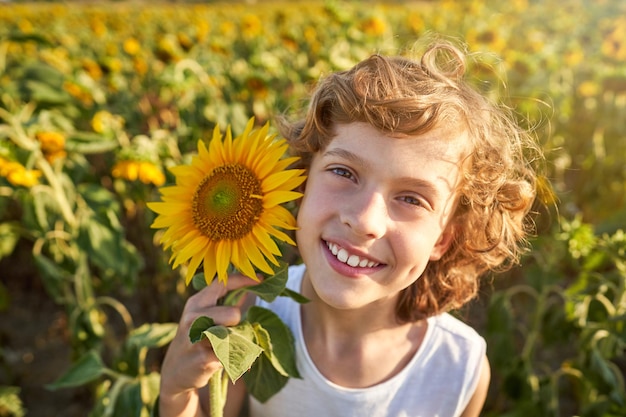 Zorgeloos kind met krullend haar glimlachend en kijkend naar de camera terwijl hij de bloeiende bloem op het zonnebloemveld op het platteland aanraakt