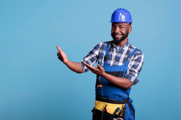 Zorgeloze bouwvakker in overall met denkbeeldig product voor de camera. Afro-Amerikaanse bouwer die advertentieruimte toont met armen aan de orde gesteld in studio-opname met blauwe achtergrond.
