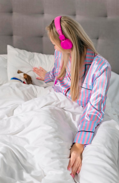 Zorgeloze dame in pyjama die naar muziek luistert op een koptelefoon terwijl ze in bed zit met haar geliefde huisdier