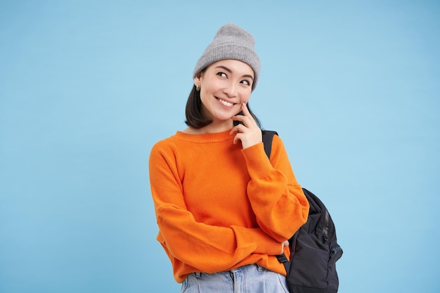 Zorgeloze mooie vrouw in streetwear houdt rugzak glimlacht en kijkt doordachte blauwe achtergrond