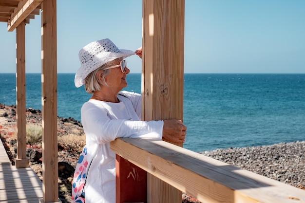 Zorgeloze oudere vrouw in een witte hoed die buiten op zee staat op een zonnige dag en naar de horizon kijkt